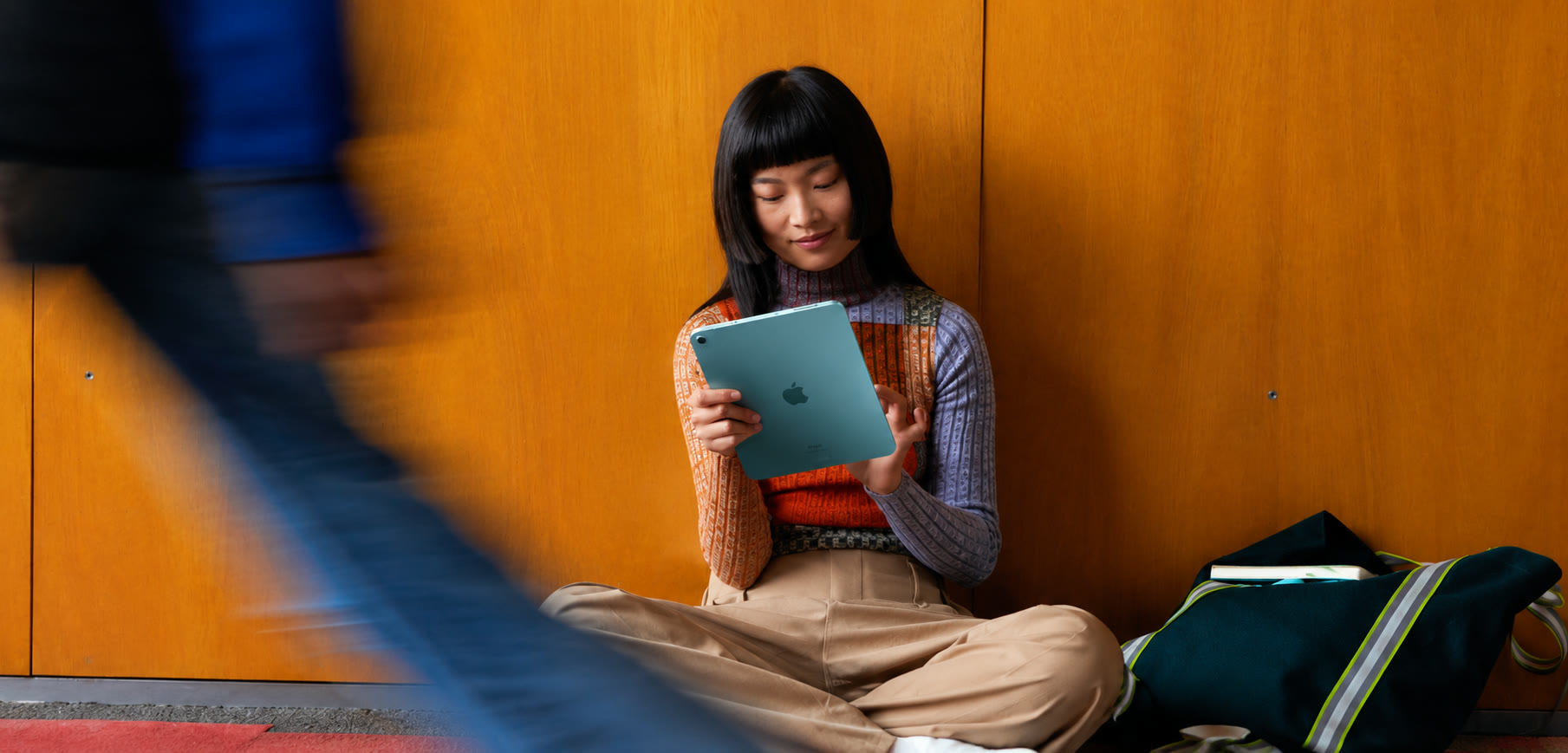 A woman sitting against a wood-panelled wall, using a teal iPad while a blurred figure walks past